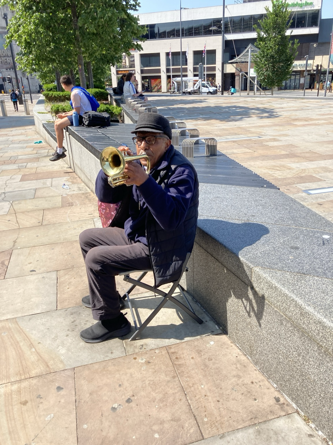 Elderly man seated in folding chair on plaza playing a cornet