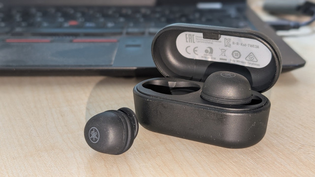 Ein Set anthrazitfahrbener In-Ear-Headsets. Ein Stöpsel steckt im offen stehenden Ladecase und ein Stöpsel liegt daneben und man kann das Yamaha-Logo erkennen. Im Hintergrund sieht man die Tastatur eines Laptops.