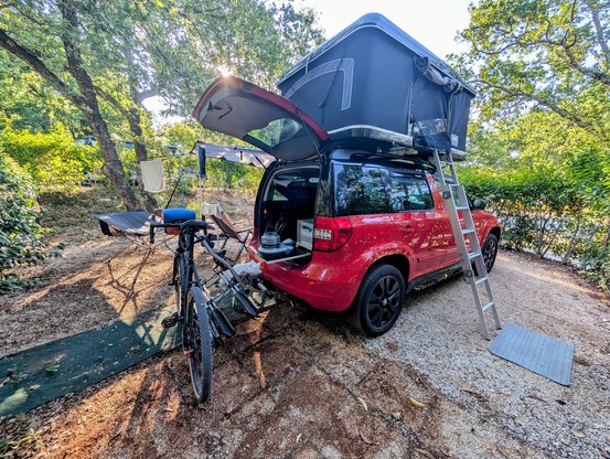 Ein roter Skoda Yeti mit einem schwarzen Dachzelt und einem Fahrradträger, an dem ein Gravelbike lehnt. Auf einem kleinen Auszug aus dem offenen Kofferraum, steht ein Gaskocher mit einem Wasserkessel.