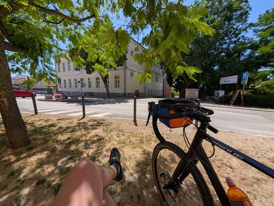 Man sieht von einem Fahrrad den vorderen Teil. Zudem die überschlagenen  Beine eines Mannes, der möglicherweise auf einer Bank sitzt, an der auch das Fahrrad lehnt. Ein Baum wirft Schatten. Davor führt eine Straße entlang. Es scheint sehr sonnig zu sein.