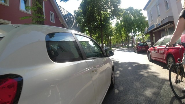 Ein weißer PKW überholt eine Radfahrerin mit zu wenig Abstand. Rechts parken PKW am Fahrbahnrand halb in Parkbuchten. Im schmalen Abstand zwischen der Radfahrerin und dem PKW ist offensichtlich noch das Fahrrad mit der mittig am Lenker montierten Kamera, welche das Bild aufgezeichnet hat.