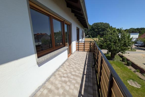 Ein von der Sonne beschienener Balkon mit beigen Fliesen. Der Blick fällt auf der schmalen Seite auf ein Feld mit einem dahinter liegenden Wald.