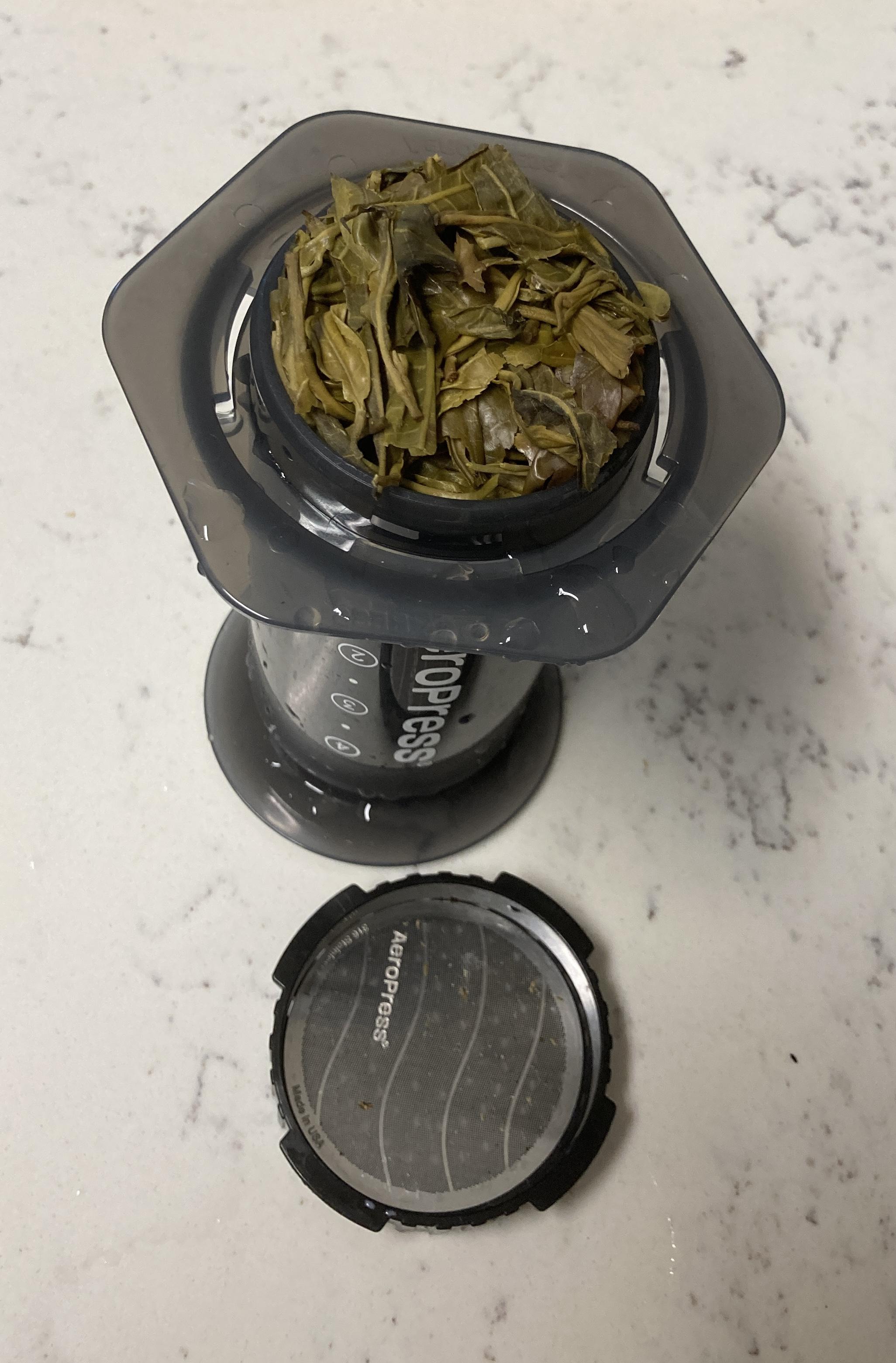 Inverted Aeropress brewing chamber with fully engaged plunger above, with green leaves threatening to spill out; Aeropress screw cap with metal filter below