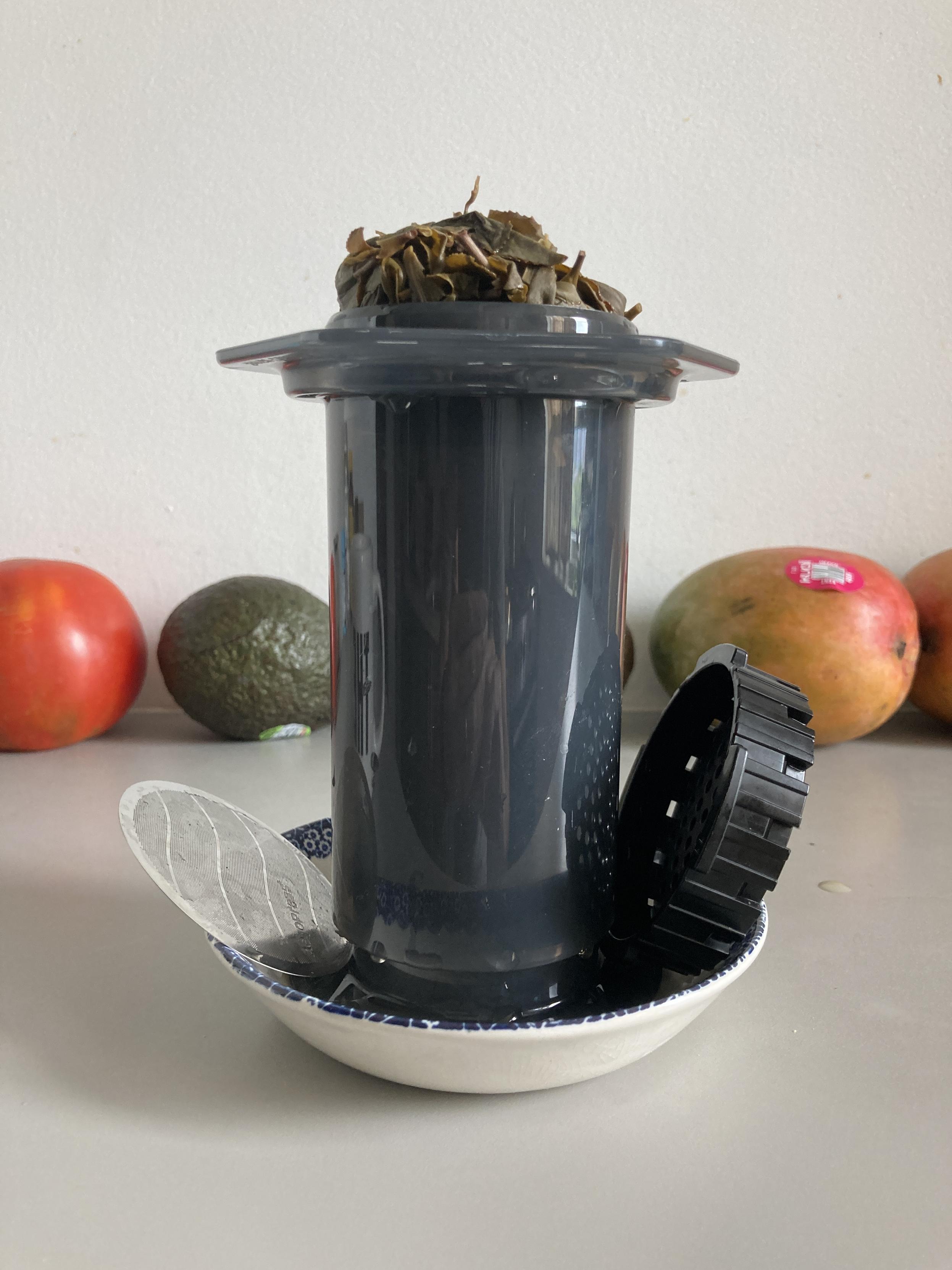 The uncapped AeroPress at eye level in a little bowl with cap and filter on the side, on a counter with a row of fruits behind it