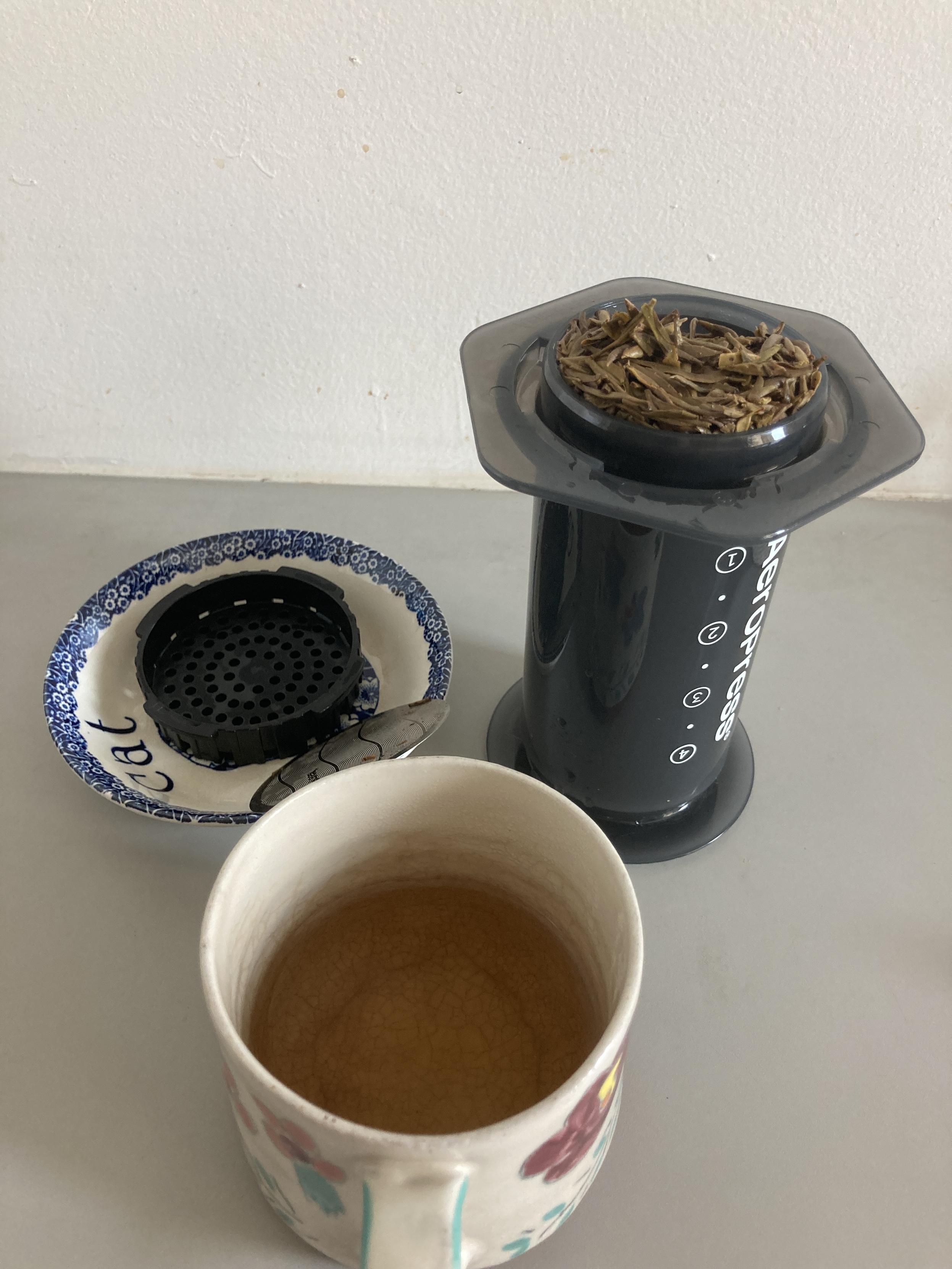On a gray counter, a hand-painted stoneware cup 1/3 full of amber tea liquor; behind it uncapped inverted Aeropress overflowing with steeped leaves and a small bowl with the cap and filter
