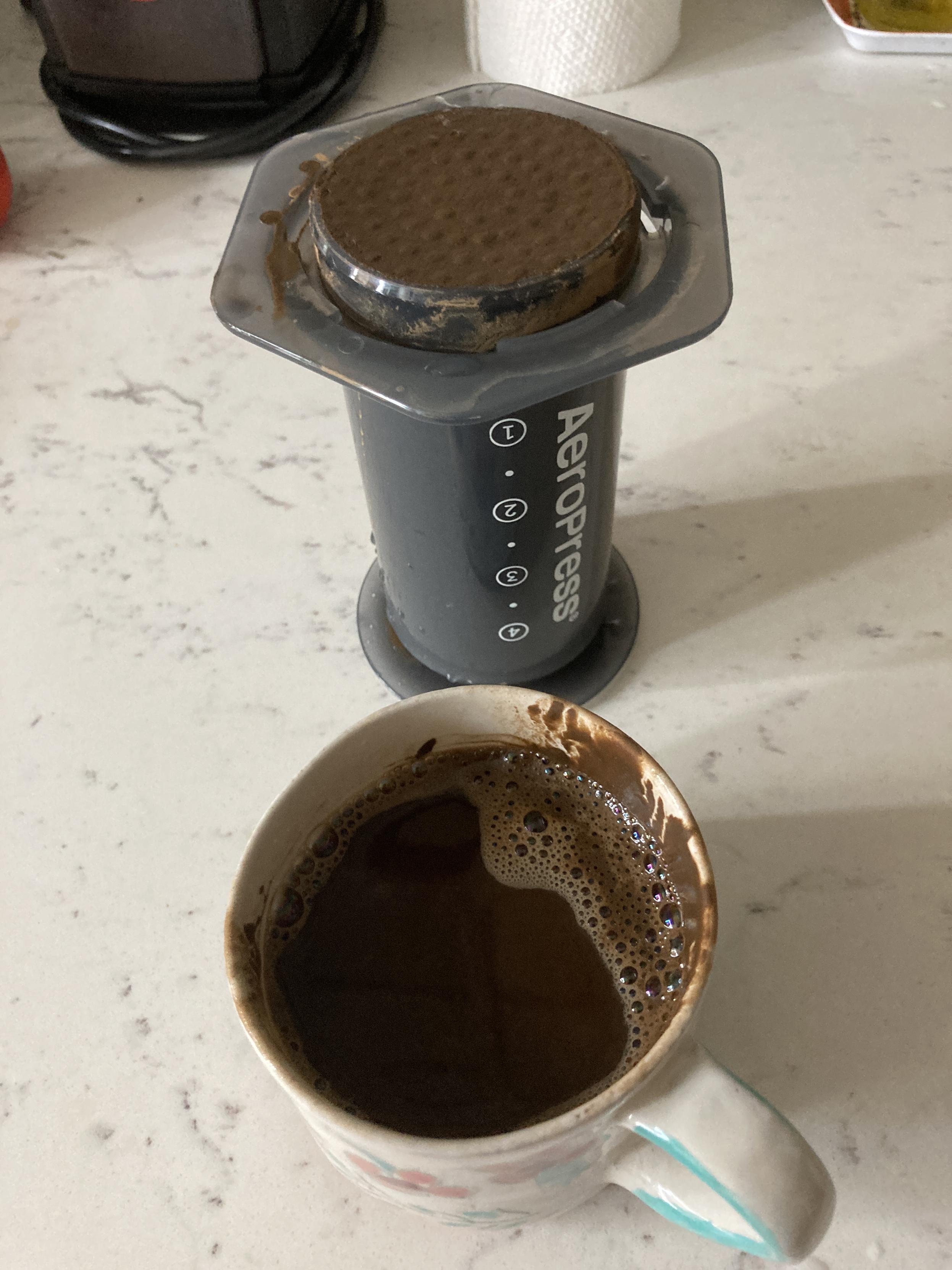 On a stone counter, a hand painted mug full of foamy brown liquid in front of an overturned uncapped Aeropress with a brown hojicha puck on top