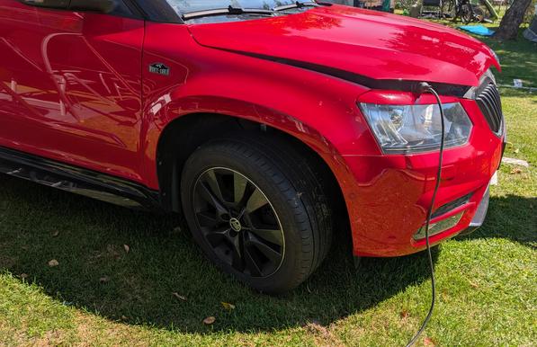 Die vordere rechte Seite eines roten Autos, dessen Motohaube einen Spalt aufsteht und ein Stromkabel in den Motorraum führt.