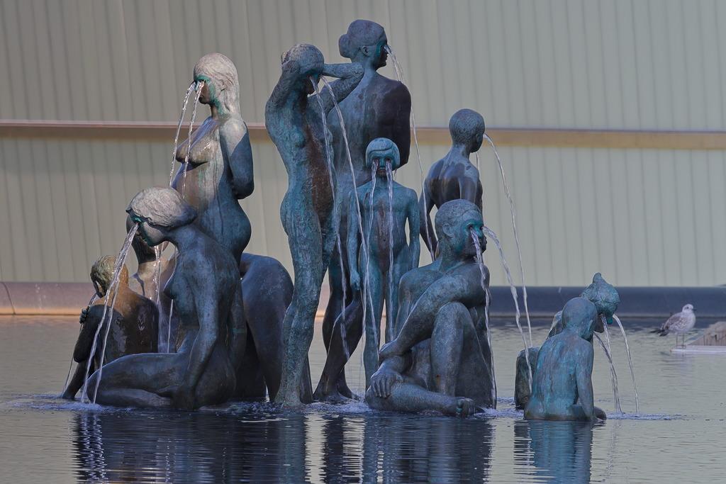 Kunstwerk Women & Children in Helsinki. Eine Gruppe von Frauen und Kindern aus Bronze, die in einer Wasserfläche dicht beieinander stehen und sitzen. Aus jedem Auge der Figuren kommt ein Wasserstrahl. Sozusagen im Strahl weinen, statt im Strahl zu kotzen.