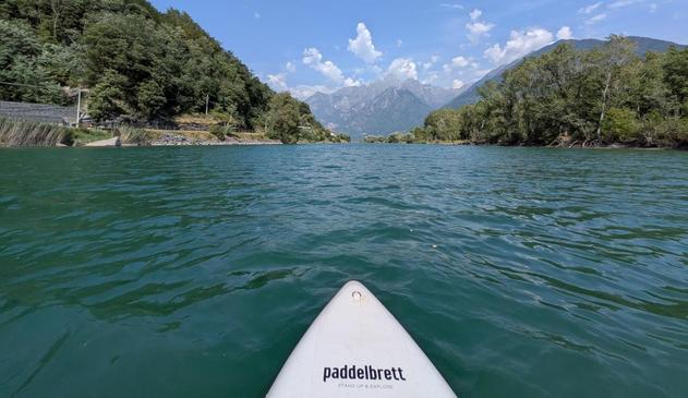 Blick über eine Wasserfläche auf von Bäumen bestandene Berge und Hügel. Im Vordergrund sieht man die Spitze eine StandUp-Boards mit der Aufschrift "Paddelbrett".