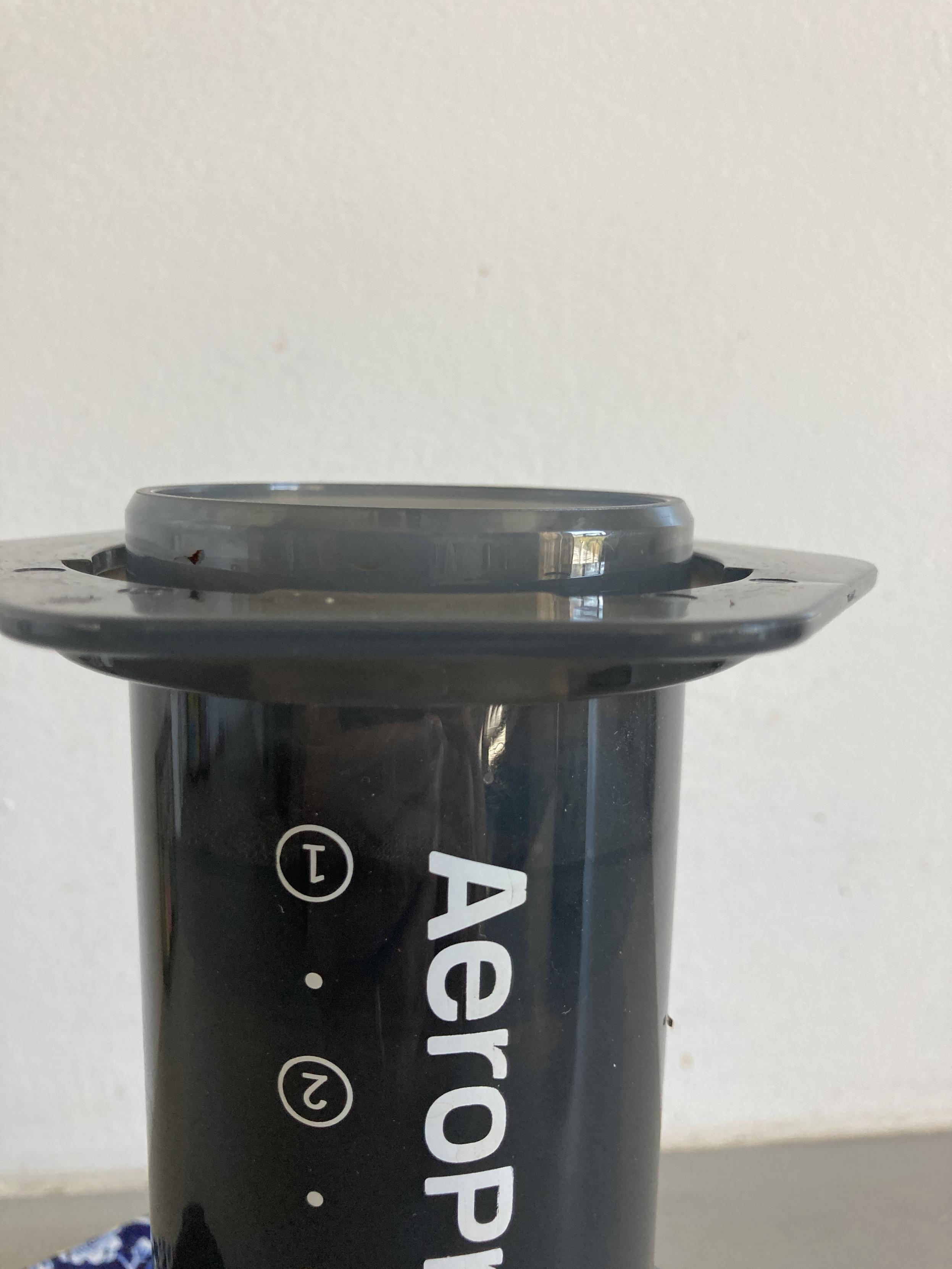 Closeup of uncapped inverted Aeropress against a wall, showing how hard it is to discern where the plunger ends