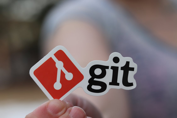 Ein noch nicht verklebter Aufkleber mit dem offiziellen Logo des Versionsverwaltungs-Tools git wird in die Kamera gehalten. Der Hintergrund ist unscharf, was den Vordergrund künsterlisch betont.

Quelle: https://www.pexels.com/de-de/foto/hand-festhalten-halten-papier-11035539/ 