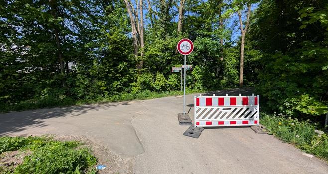Ein Absperrzaun und ein Durchfahrt-Verboten-Schild stehen halb auf einer T-Kreuzung eines Geh- und Radweges. Im Hintergrund sind Bäume und Büsche zu sehen.