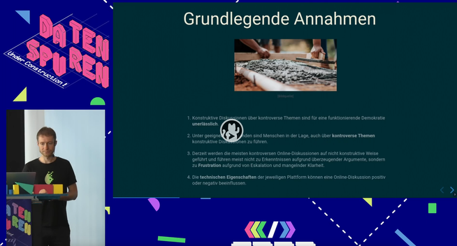 Screenshot aus dem verlinkten Video. Links: Einblendung des Referenten hinter einem Redner:innenpult; Rechts: Einblendung der Präsentation, aktuelle Folie hat die Überschrift: "Grundlegende Annahmen".