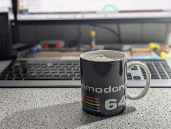 Vor einer silbernen Computertastatur steht ein schwarze Kaffeetasse mit einem Commodore Schriftzug und dem C64-Logo.