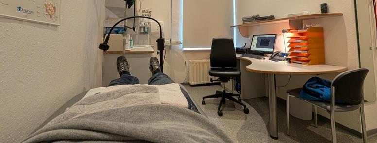 Ein Mann liegt in einem Behandlungsraum auf einer Liege. Man sieht den Köroer ab der Hüfte abwärts. im Hintergrund ein Ständer für Infusionen, an der Wand rechts ein Schreibtisch mit Stuhl und einem Computer auf der Tischplatte.