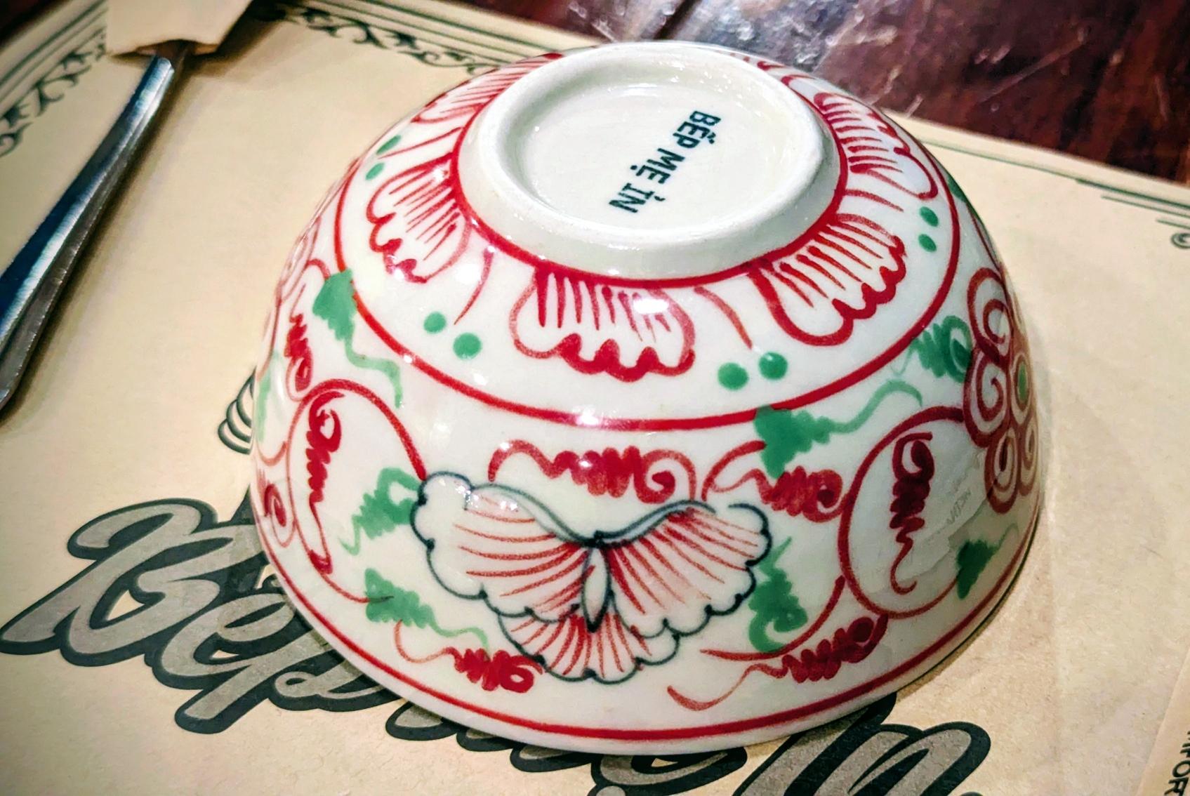 The image features a porcelain bowl in the famous Bรกt Trร ng style, named after the village of the same name near Hanoi. The bowl is decorated with a painted red and green floral pattern.