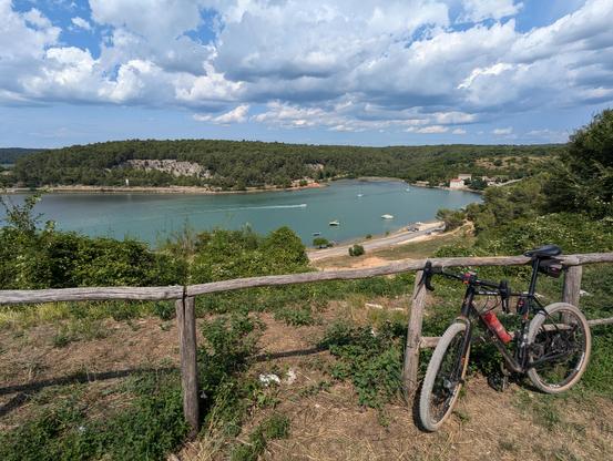 Ein Fahrrad lehnt an einem aus einfachen Rundhölzern bestehenden Zaun auf einem Hügel. Im Hintergrund sieht man eine Bucht mit grünem Wasser, welche von mit grünen Kiefern bewachsenen Hügeln umgeben ist.