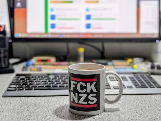 Eine Kaffeetasse mit der Aufschrift FCKNZS steht vor einer Tastatur und einem Bildschirm.