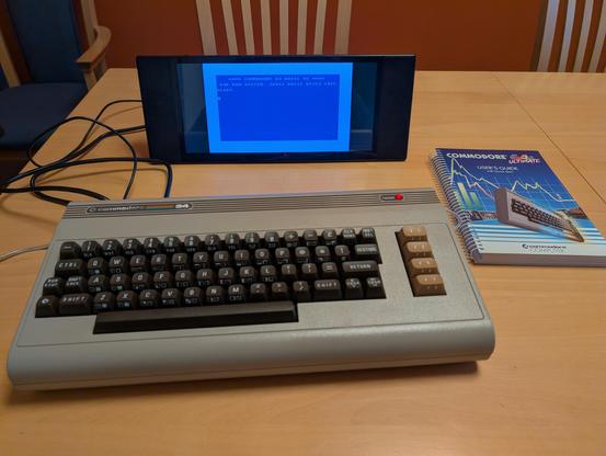 Ein Heimcomputer Commodore C64 ist an einem kleinen Flachbildschirm angeschlossen. Daneben liegt das Handbuch zum Computer.
