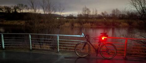 Ein Gravelbike lehnt in der Dunkelheit mit eingeschaltetem Licht an einem Geländer. Im Hintergrund fließt ein ruhiger Fluss und man sieht in der Ferne eine Hügelkette.