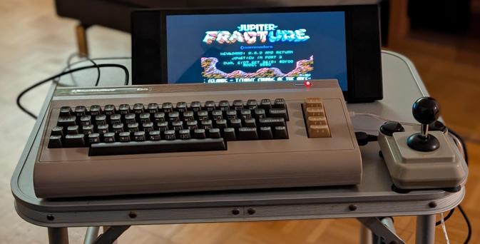 Ein C64 auf einem kleinen Campingtisch. Rechts daneben steht ein grauer Joystick und auf dem angeschlossenen Bildschirm ist der Startscreen eines Spiels zu sehen.