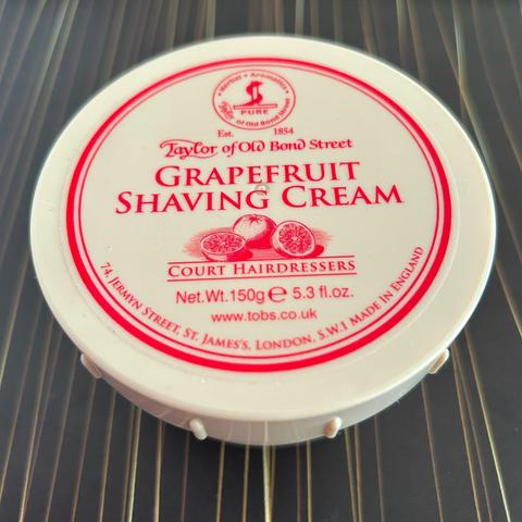 Das Bild zeigt einen runden Behälter mit Aufschrift. Es handelt sich um eine Grapefruit-Rasiercreme von Taylor of Old Bond Street. Auf dem Deckel sind der Produktname, das Gründungsjahr (1854), das Gewicht (150g / 5.3 fl.oz.) und die Adresse des Unternehmens (74 Jermyn Street, St. James's, London, S.W.1) angegeben. Außerdem steht darauf "Made in England" und eine Website-Adresse. Das Logo des Unternehmens ist ebenfalls abgebildet, zusammen mit einer Illustration von Grapefruits. Der Hintergrund…