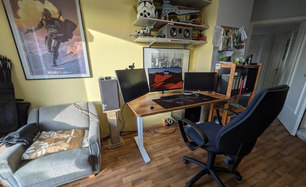 Ein Schreibtisch wird durch ein nicht sichtbares Fenster von der Sonne angestrahlt. Links im Bild ist die Hälfte eines Sessels zu sehen. Eine Lautsprecherbox steht auf einem Marmorständer und auf dem höhenverstellbaren Schreibtisch sind drei Monitore aufgebaut. An der Wand hängt ein Mandalorian-Poster und hinter den Monitoren sieht man das kitschige Poster mit den Bauarbeitern auf einem Stahlträger im New York des frühen 20. Jahrhunderts.