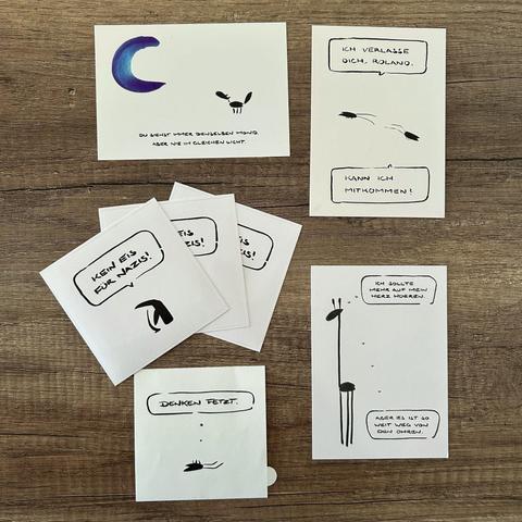 Das Bild zeigt eine Anordnung von mehreren weißen Karten und Stickern mit handgezeichneten Illustrationen und Texten auf einem hölzernen Untergrund.

Im Detail sind die Karten und Sticker auf dem Bild wie folgt beschrieben: Oben links befindet sich eine Karte mit einem blauen Halbmond und dem Text "Du siehst immer denselben Mond, aber nie im gleichen Licht". Daneben, oben rechts, ist eine Karte mit dem Text "Ich verlasse dich, Roland" in einer Sprechblase, darunter sind drei einfache Figuren, d…
