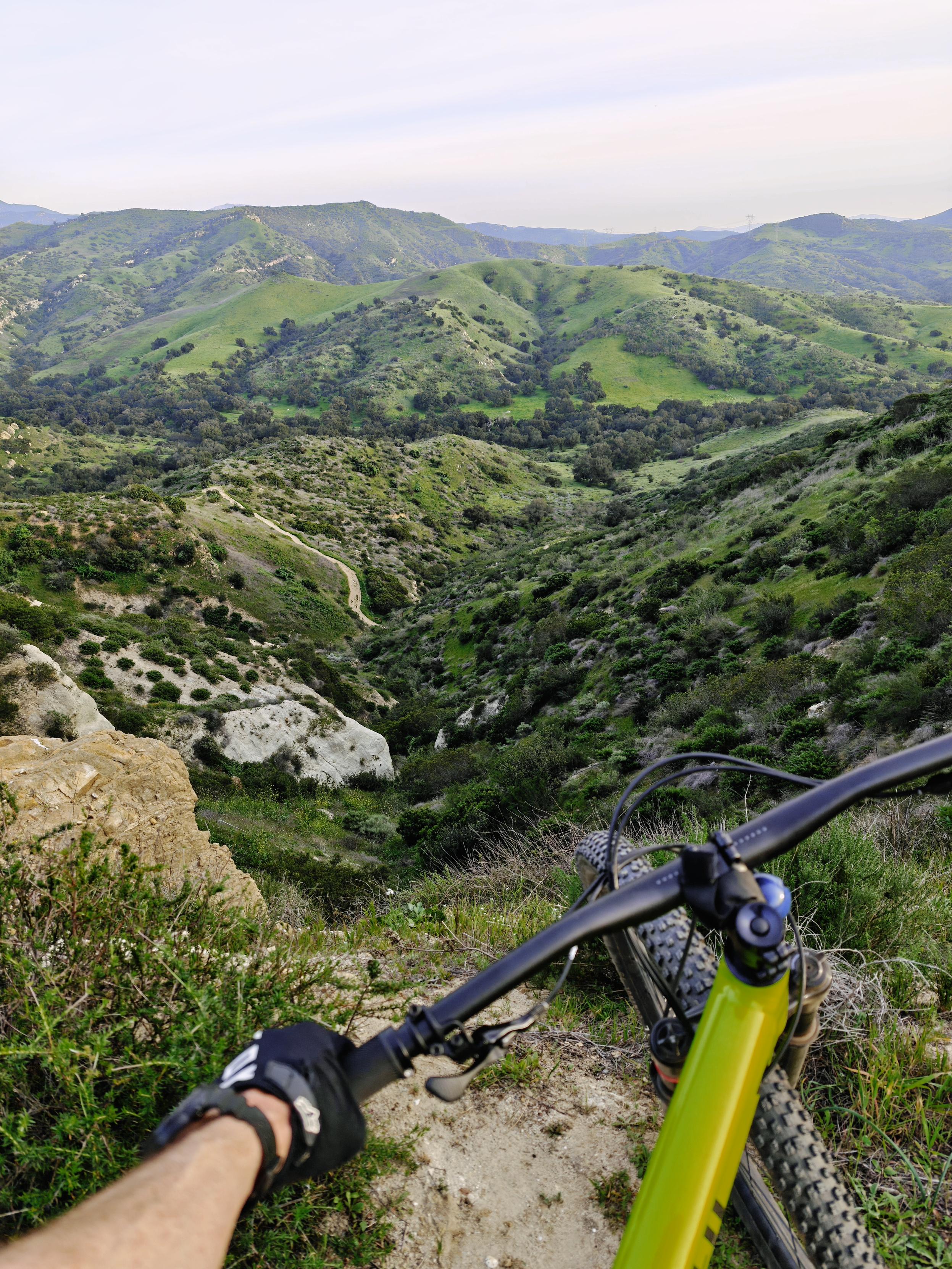 A photo taken from the top of a drop off. There are green hills to the horizon and a dirt trail that snakes around the bottom. A mountain bike and the rider's arm are in the lower foreground.