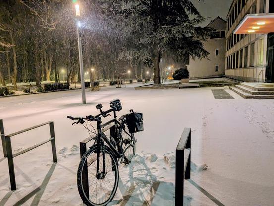 Ein Fahrrad lehnt an einem Bügel vor einem kleinen Platz. Die Szene wird con einer Straßenlampe erhellt, und man sieht Schneeflocken.