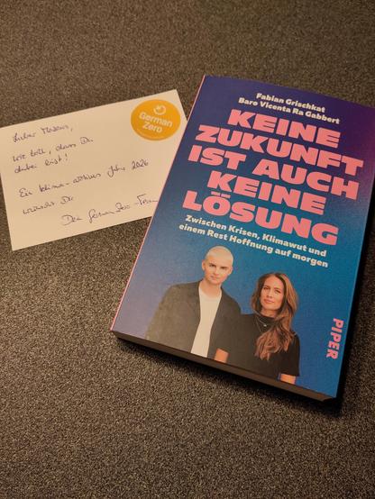 Auf einem Tisch liegen eine handschriftliche Nachricht und ein Buch nebeneinander.
Das Buch hat den Titel "Keine Zukunft ist auch keine Lösung" und trägt den Untertitel "Zwischen Krisen, Klimawut und einem Rest Hoffnung auf morgen". Die Autoren Fabian Grischkat und Bare Vicenta Ra Gabbert sind auf dem Cover abgebildet. Erschienen ist das Buch im Piper-Verlag.
Die handschriftliche Nachricht lautet "Lieber Markus, wie toll, dass du dabei bist! Ein Klima-aktives Jahr wünscht Dir Dein GermanZero-Te…