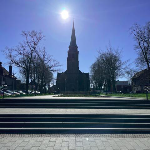 Die St. Donatus Kirche in Aachen-Brand steht im Zentrum des Bildes. Die Kirche ist von einer breiten Treppe im Vordergrund und von kahlen Bäumen umgeben. Die Sonne scheint direkt über dem Kirchturm und erzeugt einen starken Lichtkontrast, sodass die Kirche und die Bäume als dunkle Silhouetten vor dem hellen Himmel erscheinen. Links und rechts sind Wohnhäuser und weitere Gebäude zu erkennen.