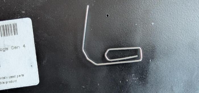 Ein kleiner Ausschnitt der Rückseite eines Lenovo Laptops. Man sieht die Ecke eines Aufklebers mit technischen Daten und eine aufgebogene Büroklammer, welche auf dem Laptop liegt. Am Ende der Büroklammer ist ein unbeschriftetes kleines Loch zu sehen.