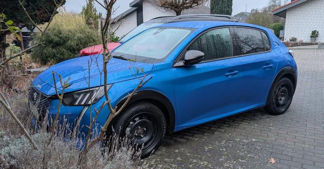 Ein blauer Peugeot 208 e-GT von schräg links vorne fotografiert. Vor der Motorhaube ist im Vordergrund eine Rosenpflanze, fast komplett versteckt hinter dem Peugeot steht ein rotes Auto. Im Hintergrund sieht man Einfamilienhäuser.