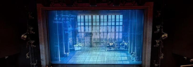 Eine Musicalbühne mit herunter gelassenem, halbtransparenten Vorhang. Hinter dem Vorhang sieht man das Bühnenbild mit einem Schreibtisch und einer kleinen Bühne mit Instrumenten.