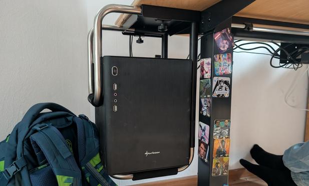 Unter einem Schreibtisch hängt ganz am linken Rand ein kleiner PCin einem aus verchromtem Rohrrahmen bestehenden Halter. Links daneben lehnt ein Schulranzen an der weißen Wand. Die sichtbare Standsäule des Schreibtisches ist mit magnetischen Star-Wars-Stickern verziert.