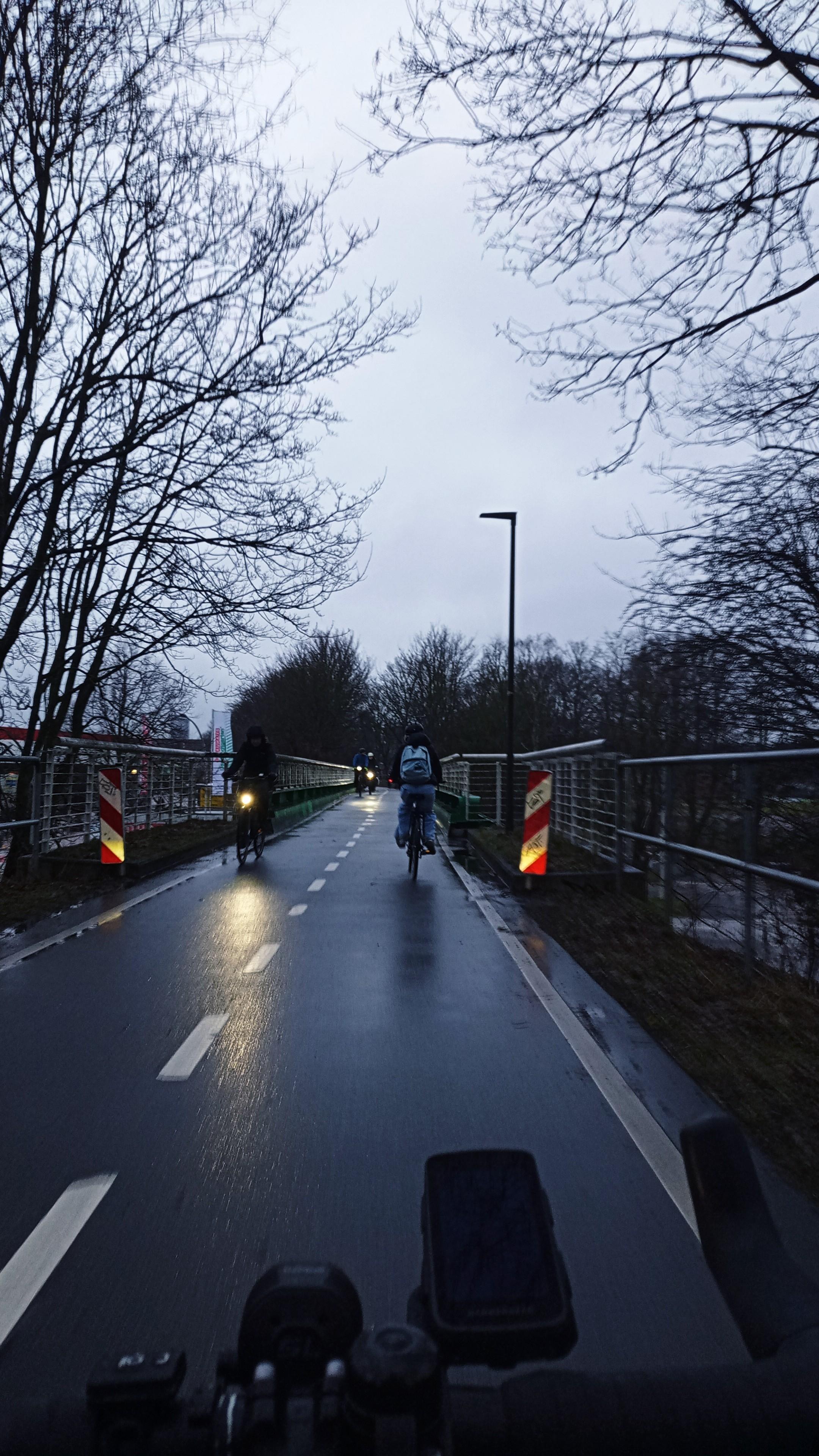 Die Veloroute 10 in Kiel im Morgengrauen. Voraus mehrere Fahrradpendler, 2 Warnbaken und eine Brücke.