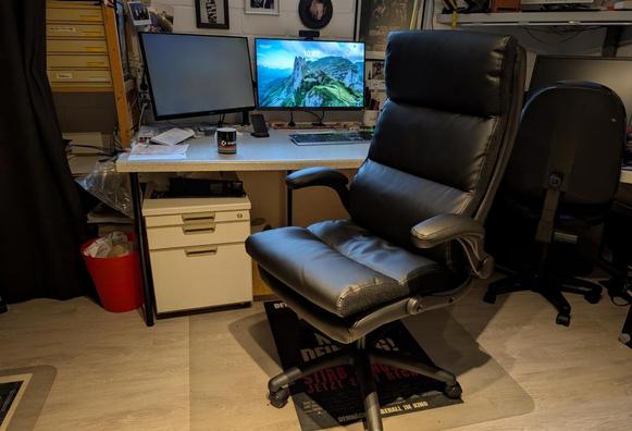 Ein schwarzer Kunstleder-Bürostuhl vor einem Schreibtisch mit zwei 27-Zoll-Monitoren. Unter dem Schreibtisch ist ein Rollcontainer zu sehen.