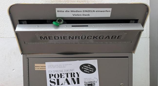 Eine Metallbox mit einer Klappe im oberen Teil. Auf der Klappe steht "Medienrückgabe". Aus der nicht ganz geschlossen Klappe schaut die Öffnung einer PET-Flasche.