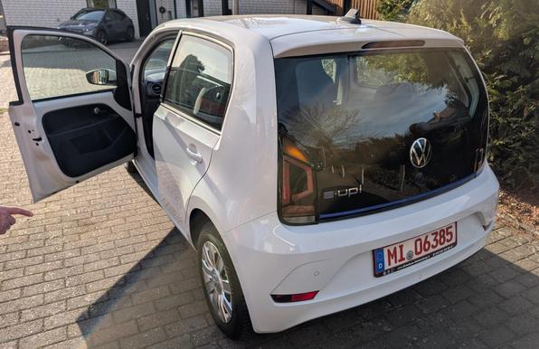 Ein von schräg hinten links fotografierter, weiß Volkswagen e-Up. Die vordere linke Tür steht offen. Es ist ein rotes Nummernschild montiert.
