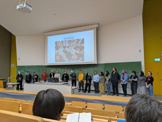 Our team of organizers and student assistants on stage waving a good bye. 18 people in a lecture hall, the photo is shot from the seating area.