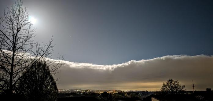 Ein Wolkenband ist klar abgegrenzt horizontal in der Mitte des Bildes zu sehen. Links scheint der Vollmond sehr grell, unter den Wolken sieht man neben Bäumen in der Ferne auch die Lichter einer Stadt. Über der Wolkengrenze ist der dunkelblaue Himmel zu sehen!