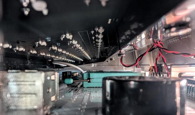 Blick von sehr flach über die Platine eines Computers. Es sind einige gesockelte Aufbauten zu sehen. Man kann aufgrund des flachen Winkels Leiterbahnen auf der Platine erahnen. Gleichzeitig blickt man von unten auf die Unterseite einer Tastatur. Rechts im Bild sind einige rote und schwarze Drähte zu sehen.