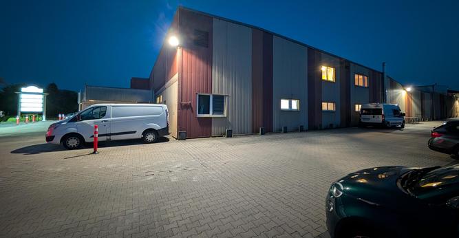 Parkplatz vor einer Gewerbehalle im Dunklen. An der Ecke der Halle ist ein Strahler montiert, der den Parkplatz erleuchtet. Rechts sieht man die Front eine Fiat 500e.