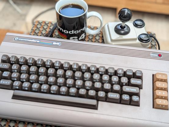 Ein C64 Ultimate auf einem Hocker. Dahinter eine gefüllte Kaffeetasse mit der Aufschrift Commodore C64 und ein ArcardeR Joystick.