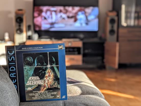 Die Hülle einer Laserdisc von 1993. Der Film ist "Krieg der Sterne". Im Hintergrund ist unscharf ein Fernseher mit dem Film zu sehen.