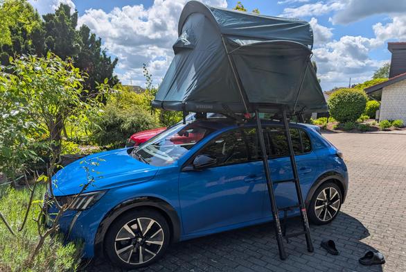 Ein blauer Peugeot E 208 GT steht auf einem Parkplatz mit einem Garten im Hintergrund. Auf dem Dach des Peugeot ist ein grünes Dachzelt "Groenberg Weekend Comfort" montiert. Man schaut von schräg vorne auf die Fahrerseite des Fahrzeugs. An die Seite des Dachzeltes ist eine Ausziehleiter angelehnt. Am eigentlich blauen Himmel ziehen reichlich weiße Wolken vorbei.