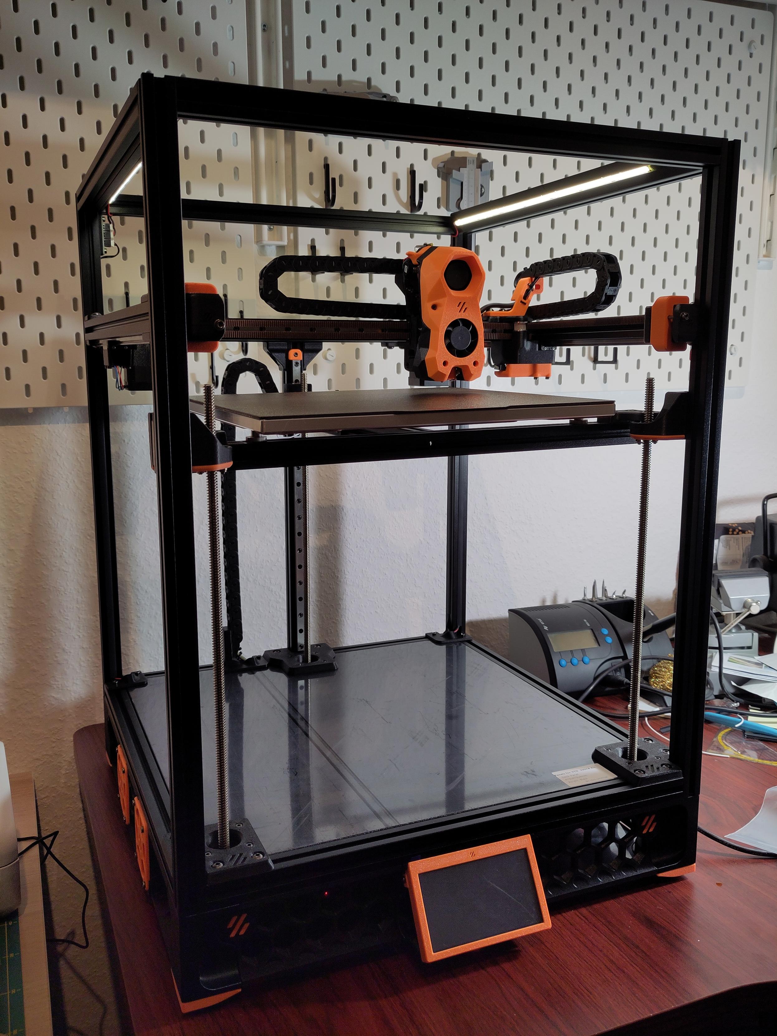 An unfinished Voron Trident, which is turned on and running, with illuminated LED strips on top.

The printer is still missing the panels and doors.