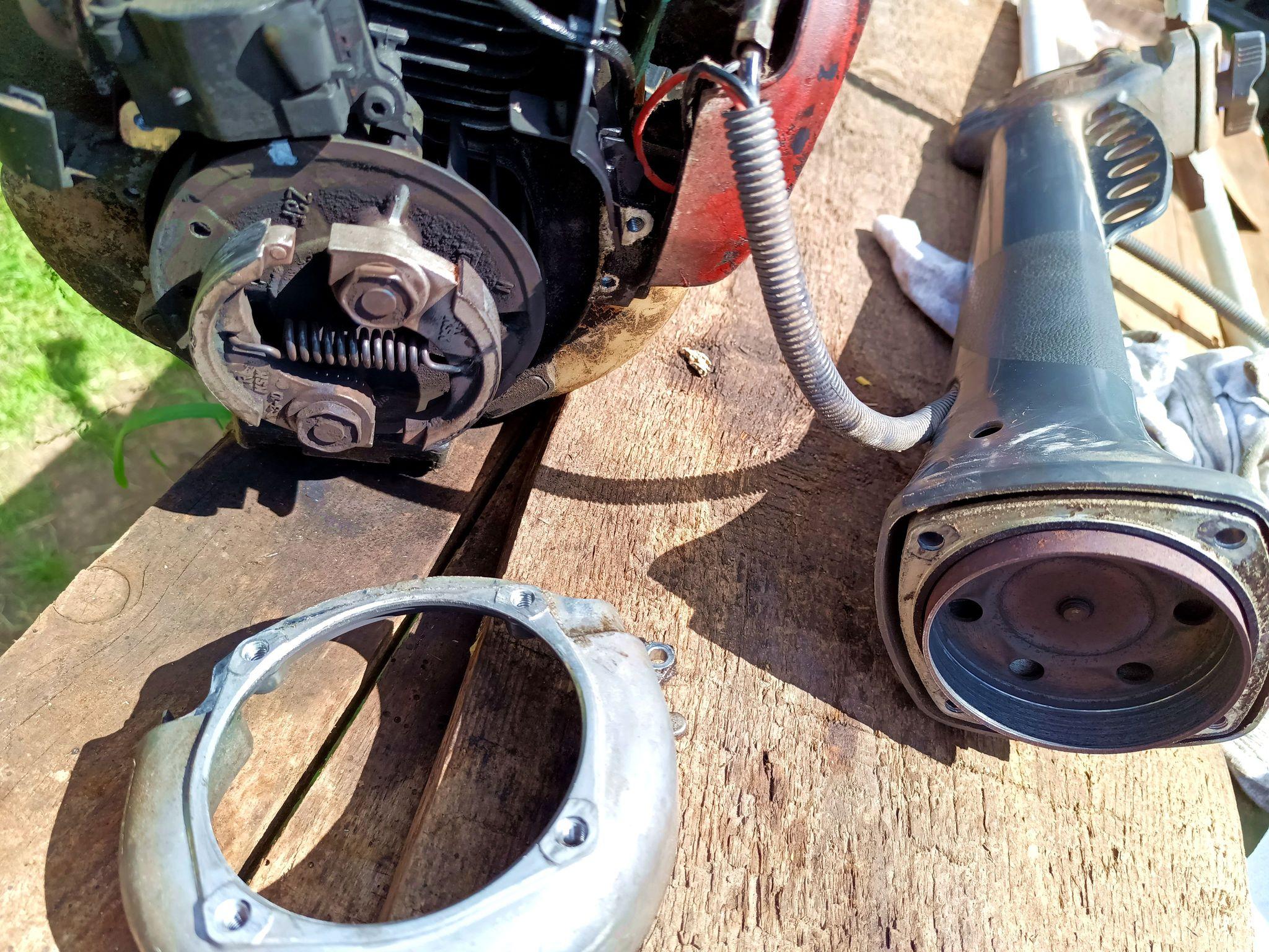 The head and the strimmer shaft are separated but still attached by the control cable bundle. The clutch housing is off too, exposing the flywheel with the centrifugal clutch attached to it that engages with the clutch bell on the shaft. There's white marker paint on the flywheel now.
