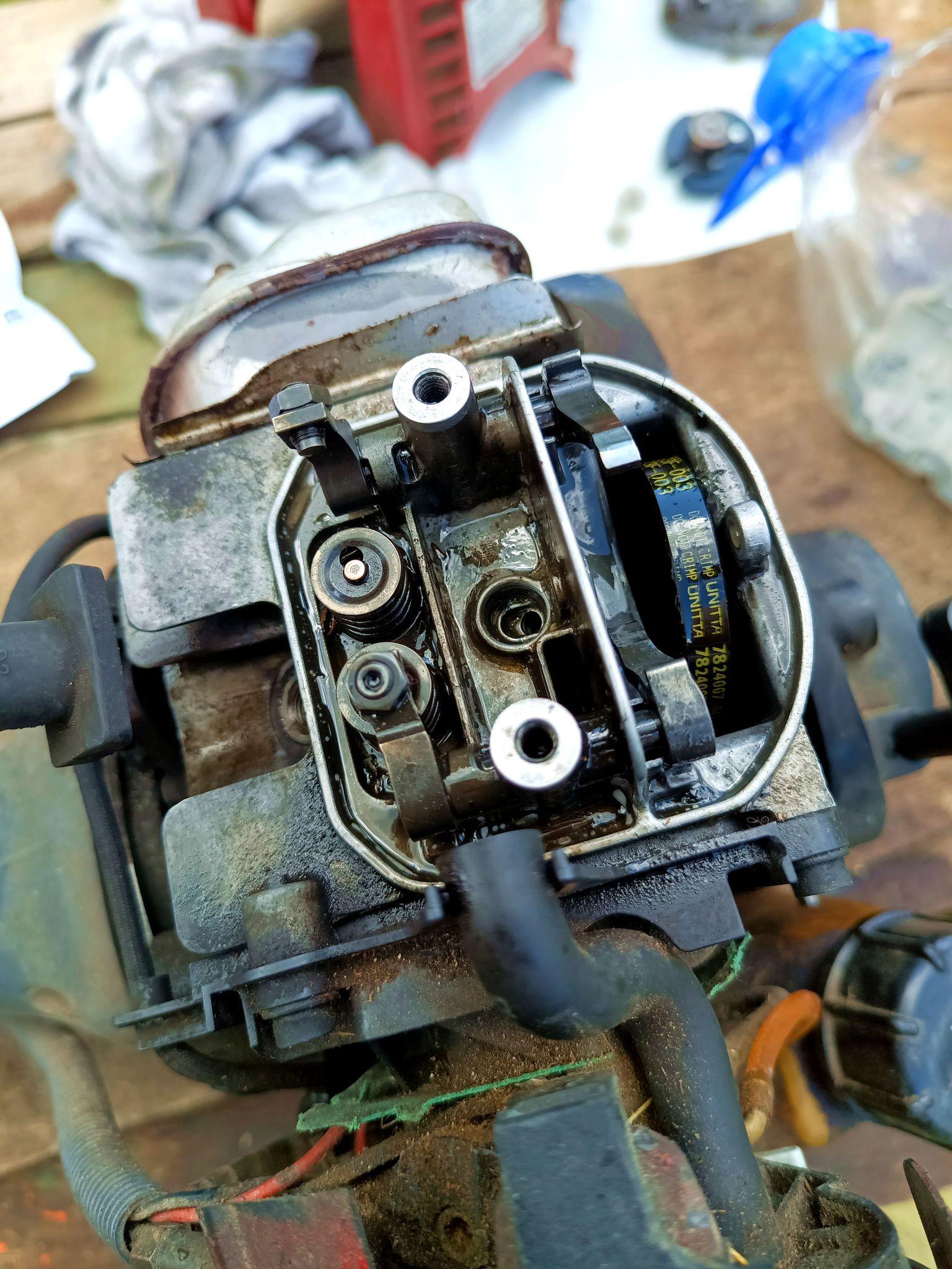 Open valve cover of a tiny 4-stroke petrol engine. On the left the intake and exhaust valve, the latter being flipped upwards. The timing pulley is missing but the timing belt is present. There's oil and some dirt in there, too.